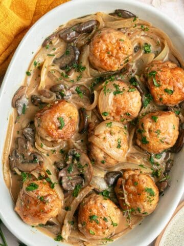 One Pan Chicken Marsala Meatballs in a creamy mushroom and onion sauce, garnished with parsley, served in a white oval dish. A yellow cloth napkin and fresh herbs are partially visible beside this gluten free meal.
