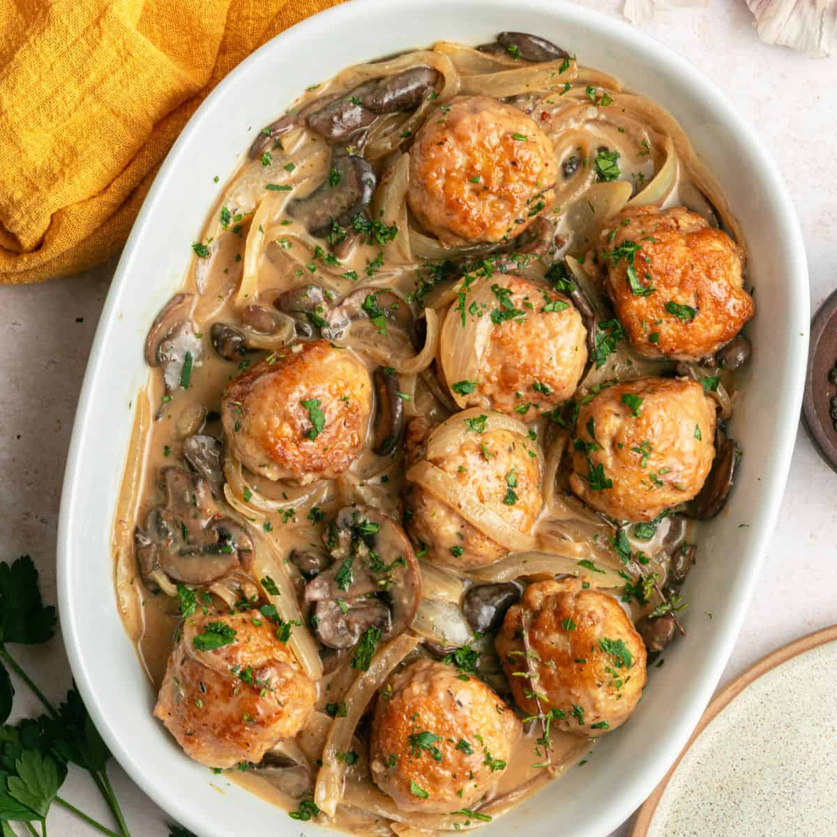 One Pan Chicken Marsala Meatballs in a creamy mushroom and onion sauce, garnished with parsley, served in a white oval dish. A yellow cloth napkin and fresh herbs are partially visible beside this gluten free meal.