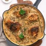 A skillet filled with four browned chicken thighs in a creamy mushroom sauce, inspired by Chicken Marsala, garnished with fresh parsley and herbs, sits on a wooden board with a small dish of ground pepper nearby.