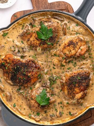 A skillet filled with four browned chicken thighs in a creamy mushroom sauce, inspired by Chicken Marsala, garnished with fresh parsley and herbs, sits on a wooden board with a small dish of ground pepper nearby.