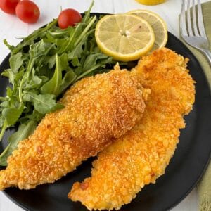 A black plate with two crispy Gluten-Free Chicken Milanese fillets, a handful of fresh arugula, two slices of lemon, and cherry tomatoes in the background. A fork and napkin are beside the plate.