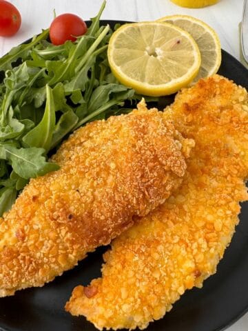 A black plate with two crispy Gluten-Free Chicken Milanese fillets, a handful of fresh arugula, two slices of lemon, and cherry tomatoes in the background. A fork and napkin are beside the plate.