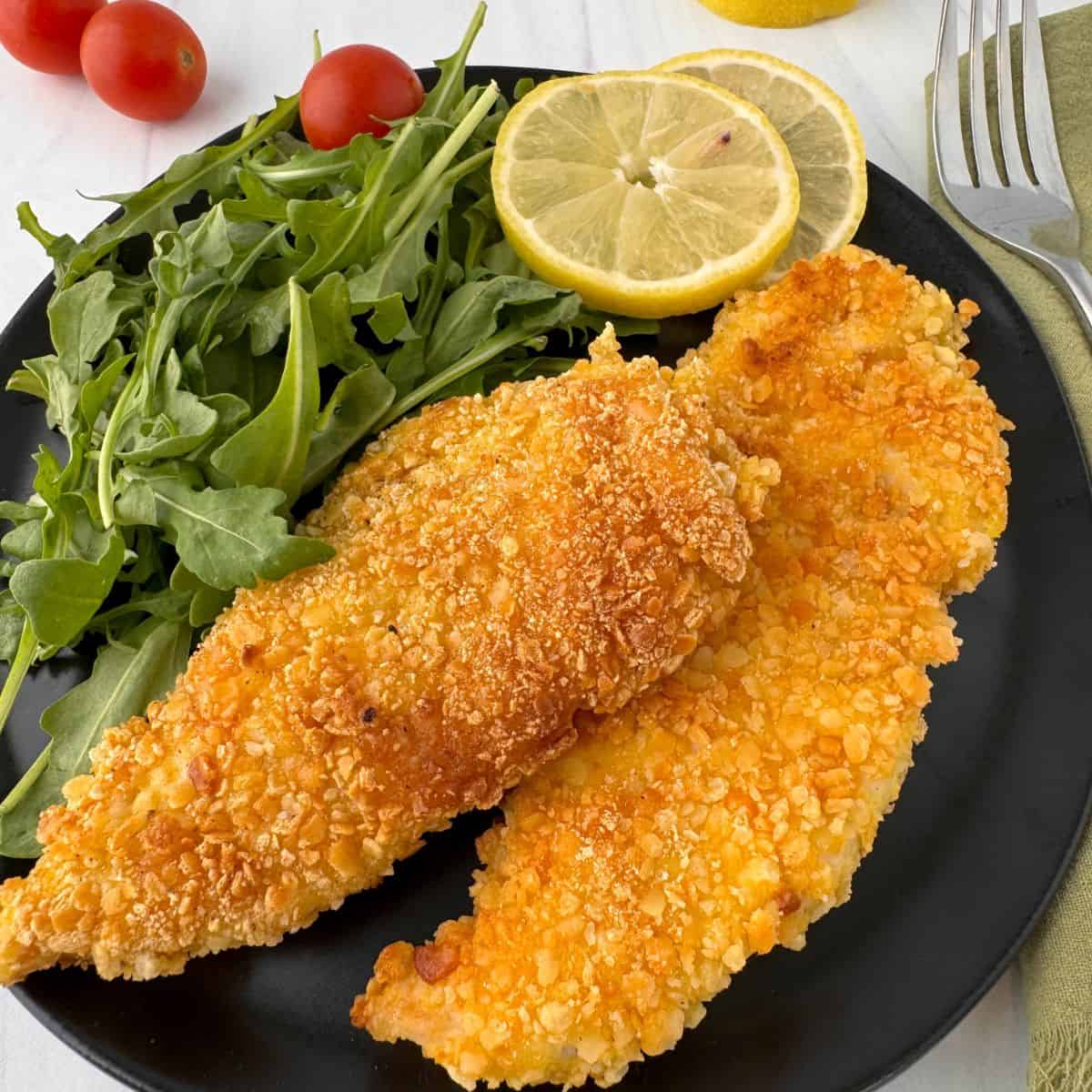 A black plate with two crispy Gluten-Free Chicken Milanese fillets, a handful of fresh arugula, two slices of lemon, and cherry tomatoes in the background. A fork and napkin are beside the plate.