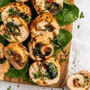 Sliced chicken roulade with spinach, sundried tomatoes, and herbs, arranged on fresh spinach leaves atop a wooden board, garnished with chopped parsley and a drizzle of balsamic glaze.