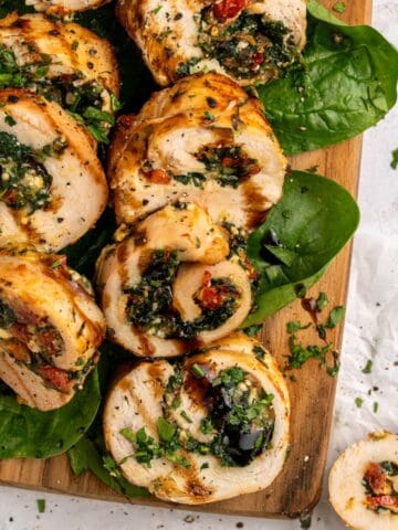 Sliced chicken roulade with spinach, sundried tomatoes, and herbs, arranged on fresh spinach leaves atop a wooden board, garnished with chopped parsley and a drizzle of balsamic glaze.