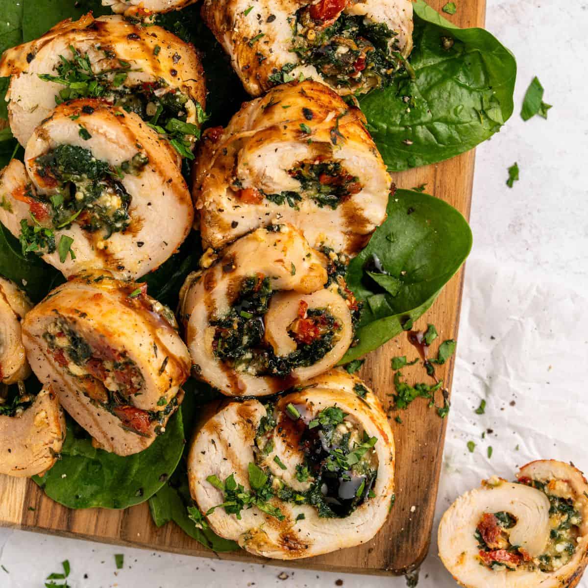 Sliced chicken roulade with spinach, sundried tomatoes, and herbs, arranged on fresh spinach leaves atop a wooden board, garnished with chopped parsley and a drizzle of balsamic glaze.