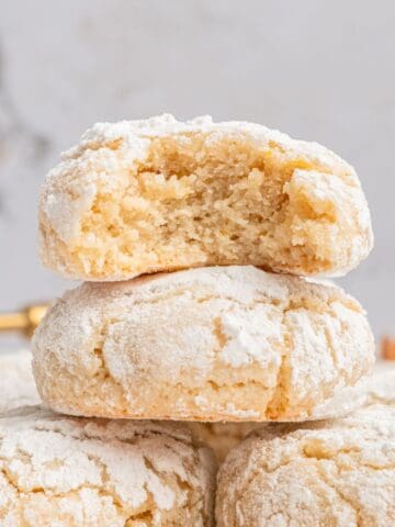 Two Italian almond cookies with almond flour, dusted in powdered sugar, are stacked; the top cookie has a bite missing, exposing its soft, crumbly interior. The background is light and blurred.