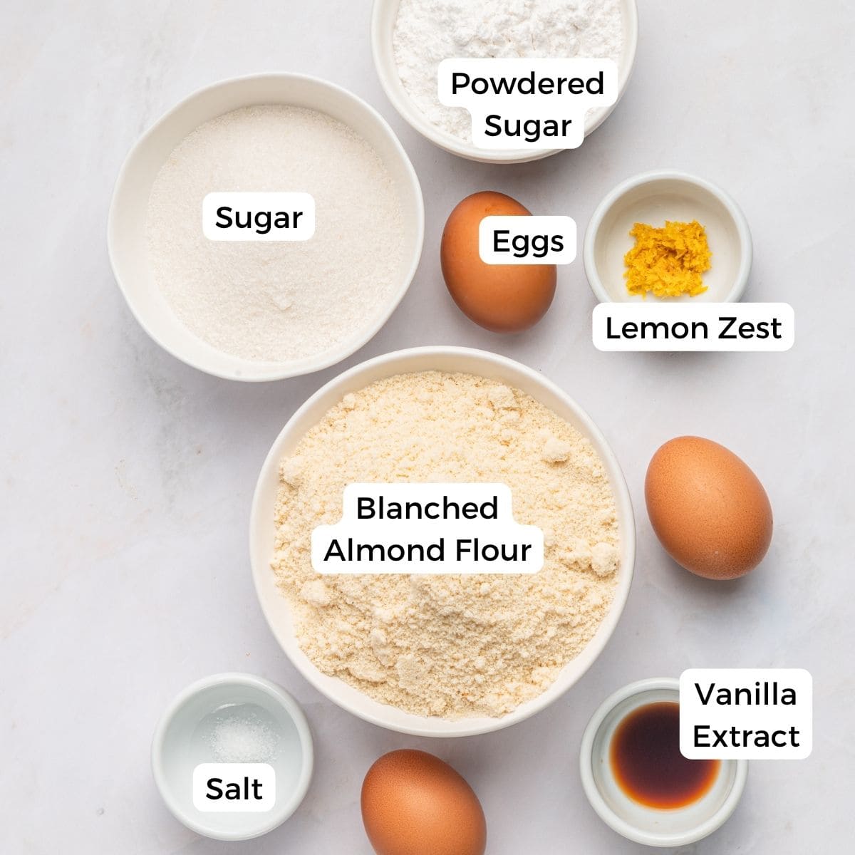 Bowls and ingredients labeled: powdered sugar, sugar, eggs, lemon zest, blanched almond flour, vanilla extract, and salt are arranged on a white surface—perfect for making Italian almond cookies with almond flour.