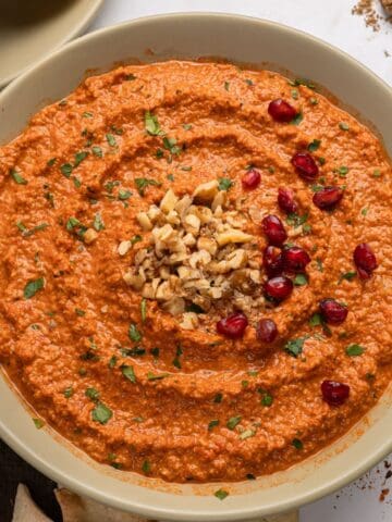 A bowl of muhammara dip, made from a classic muhammara recipe, garnished with chopped walnuts, parsley, and pomegranate seeds, displayed on a light-colored plate against a white background.