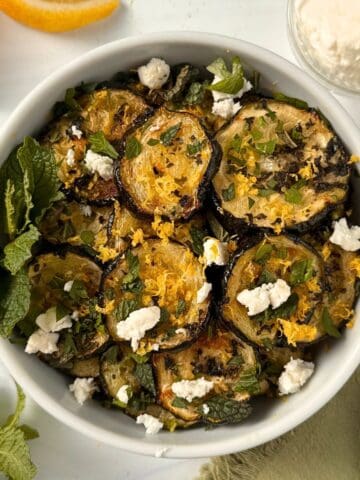 A white bowl filled with roasted zucchini slices, garnished with crumbled cheese, fresh mint, chopped herbs, and lemon zest. Orange wedges and a small dish of cheese rest nearby on a white surface.