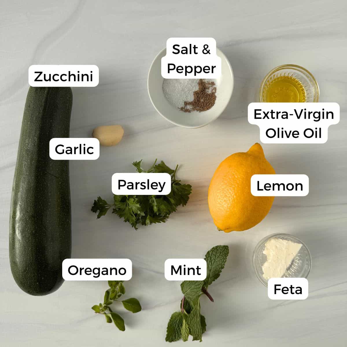 Roasted zucchini slices, garlic clove, parsley, oregano, mint, lemon, feta cheese, and extra-virgin olive oil are arranged on a light gray surface with labels for each ingredient, alongside a bowl of salt and pepper.