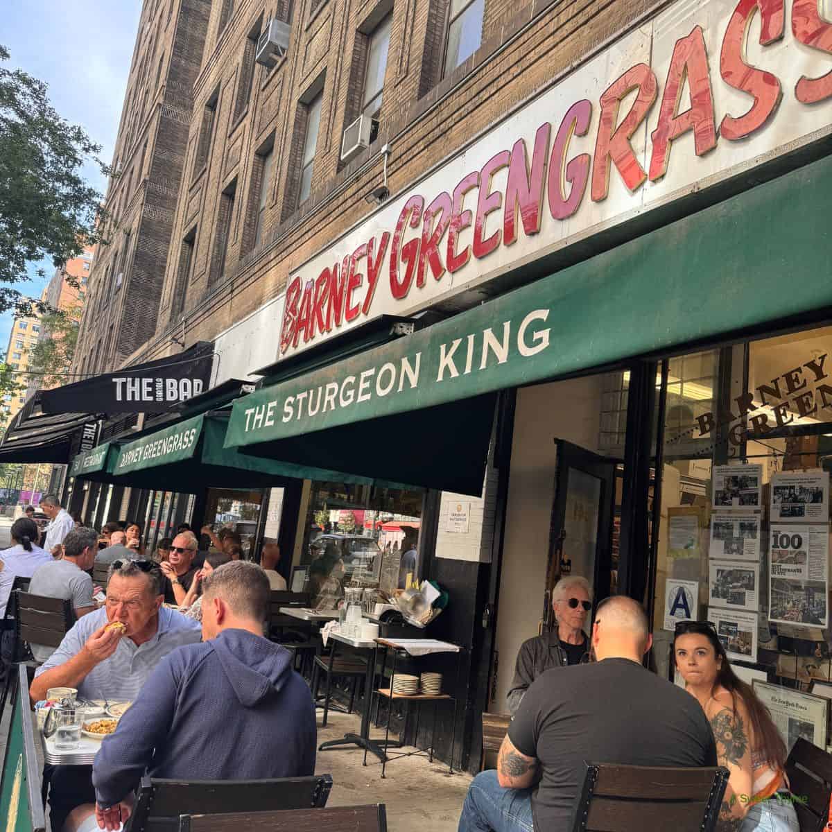 People dine outdoors at Barney Greengrass, The Sturgeon King, a historic deli with green awnings on a bustling city sidewalk—a scene straight out of The Sunday Thymes, capturing lively patrons eating and chatting.
