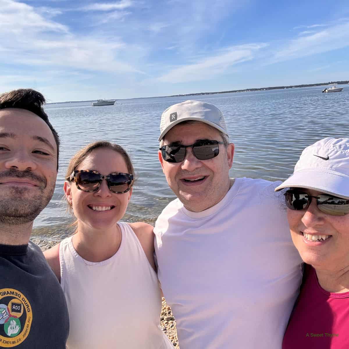 Four people smiling and taking a selfie together on a sunny day at the beach, with calm water and boats in the background—capturing memories perfect for The Sunday Thymes.