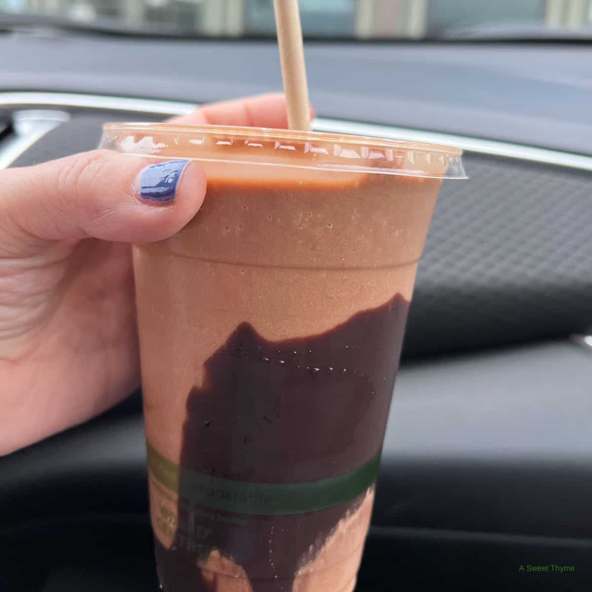 A hand with blue nail polish holds a chocolate smoothie in a clear cup with a straw, featuring swirls of chocolate syrup. Against the backdrop of a car dashboard, this sweet moment is brought to you by The Sunday Thymes.