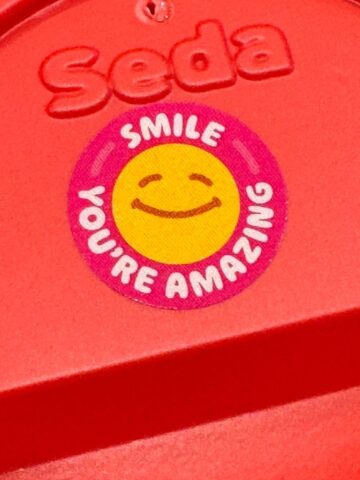 A close-up of a red surface with the embossed word Seda and a colorful sticker showing a smiling yellow face. The sticker, from The Sunday Thymes, reads, SMILE YOURE AMAZING in white letters on a pink background.