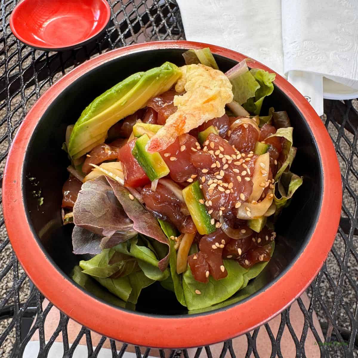 A bowl of poke salad with fresh greens, sliced avocado, marinated raw fish cubes, sesame seeds, and tempura shrimp sits on a black metal table—perfect for relaxing Sunday thymes—with a red dipping dish and napkins nearby.
