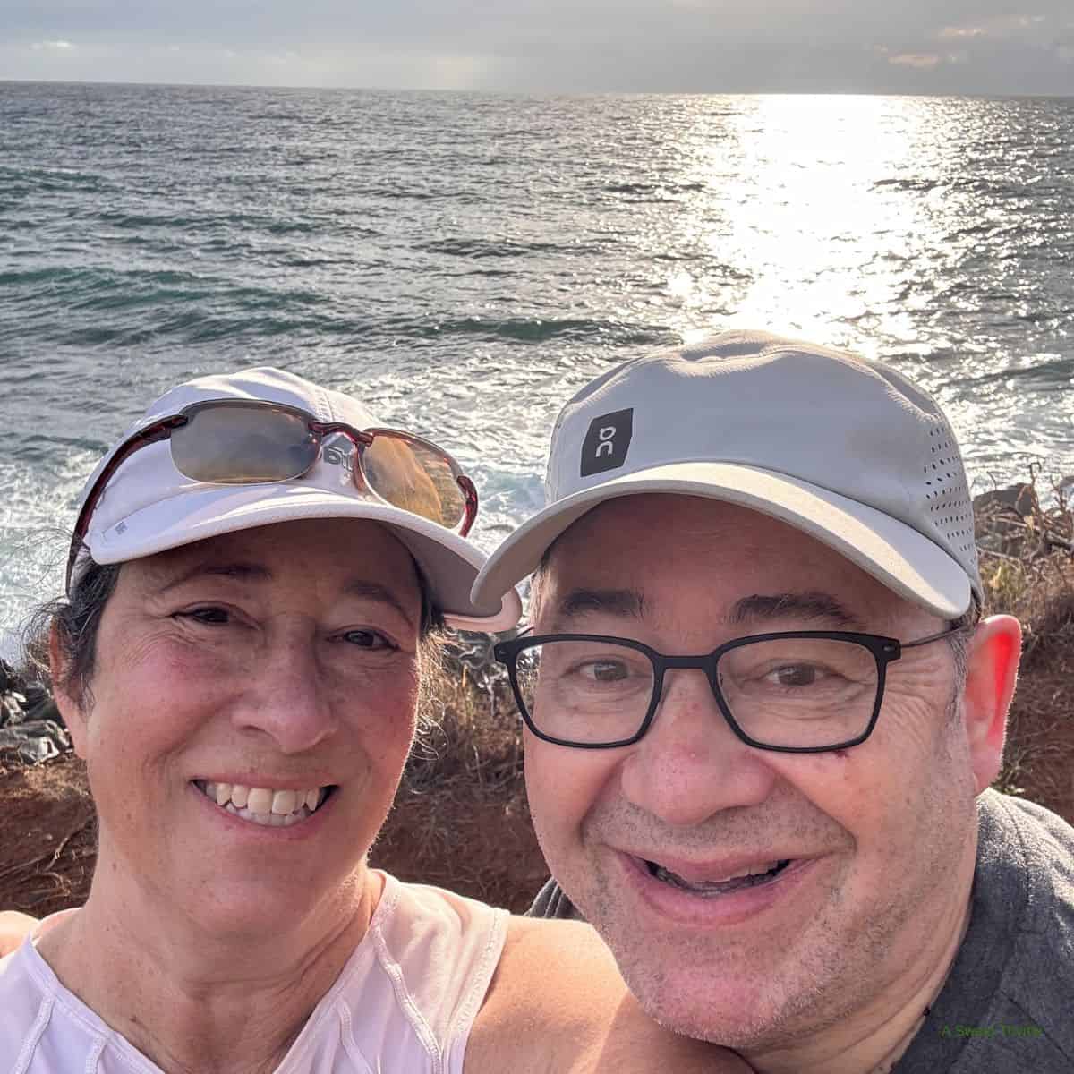 A smiling couple takes a selfie near the ocean, sunlight reflecting off the water behind them. Both wear casual clothing and hats, perhaps enjoying a relaxing morning with the Sunday Thymes as the rocky shoreline stretches out in the background.