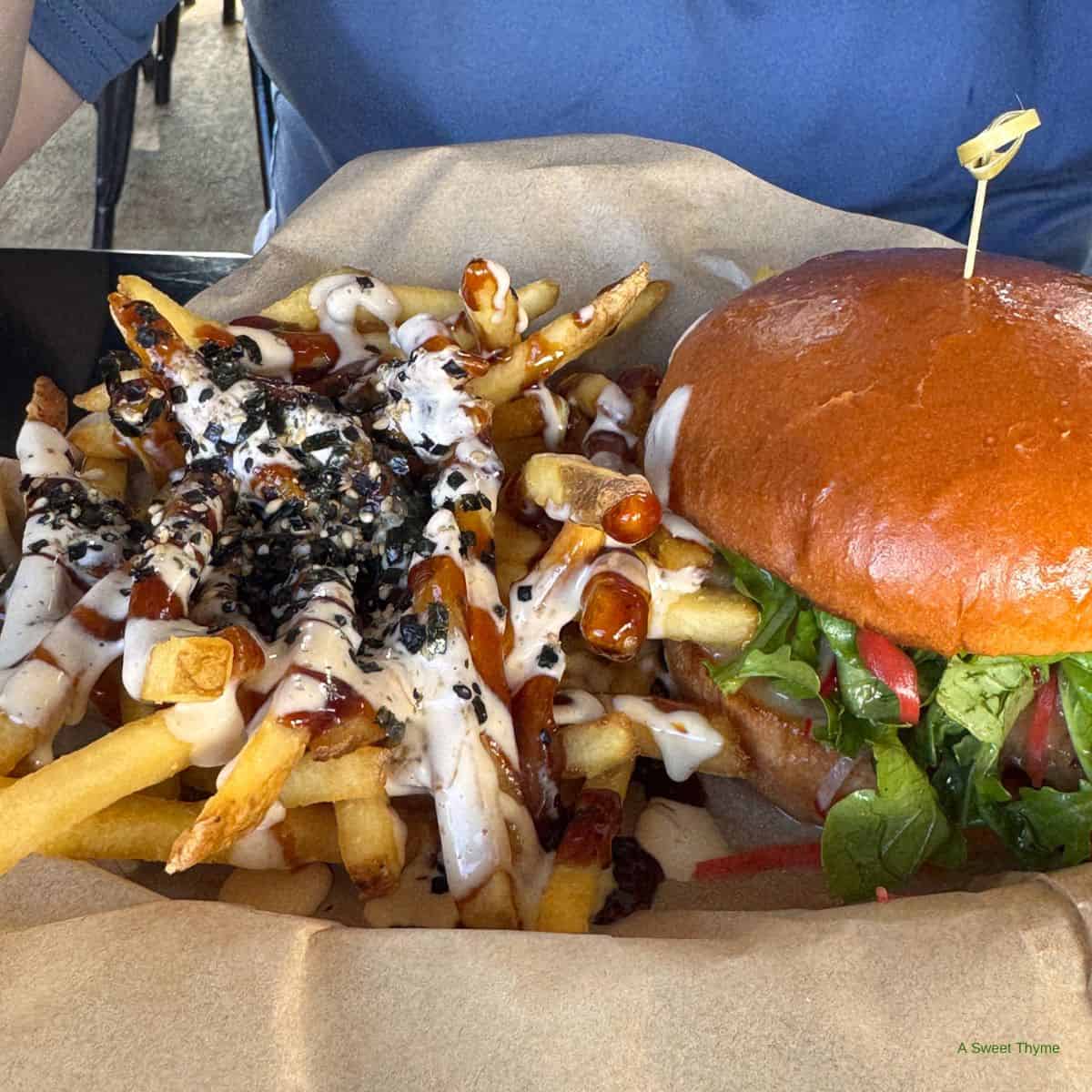 A close-up of a meal at The Sunday Thymes: a burger filled with greens, tomato, and sauce in a glossy bun, next to fries topped with white and dark sauces plus black sesame seeds on brown parchment paper.