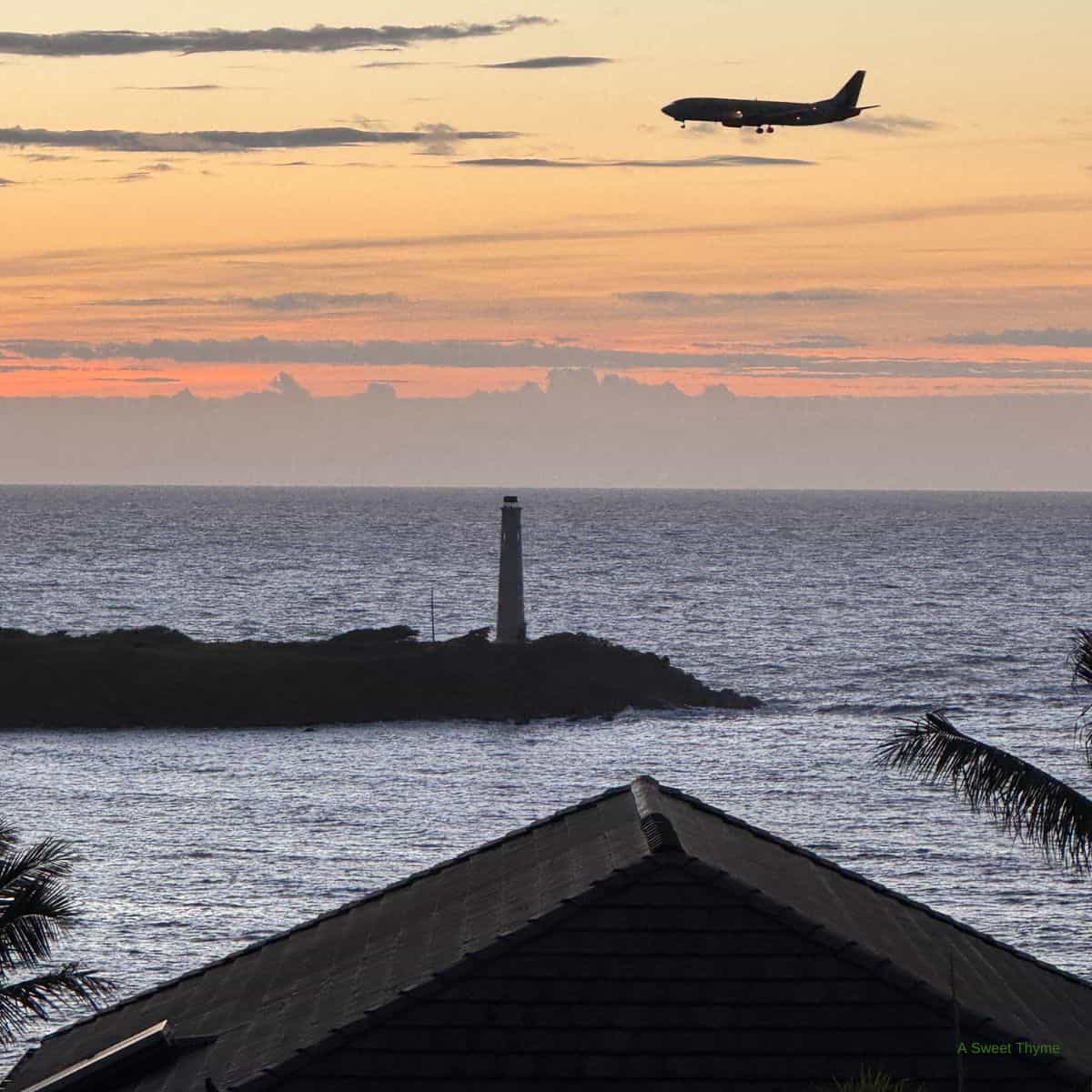 An airplane flies over the ocean at sunset, with an orange and pink sky. Below, a lighthouse stands on a small island, while Sunday Thymes rooftops and palm leaves are visible in the foreground.