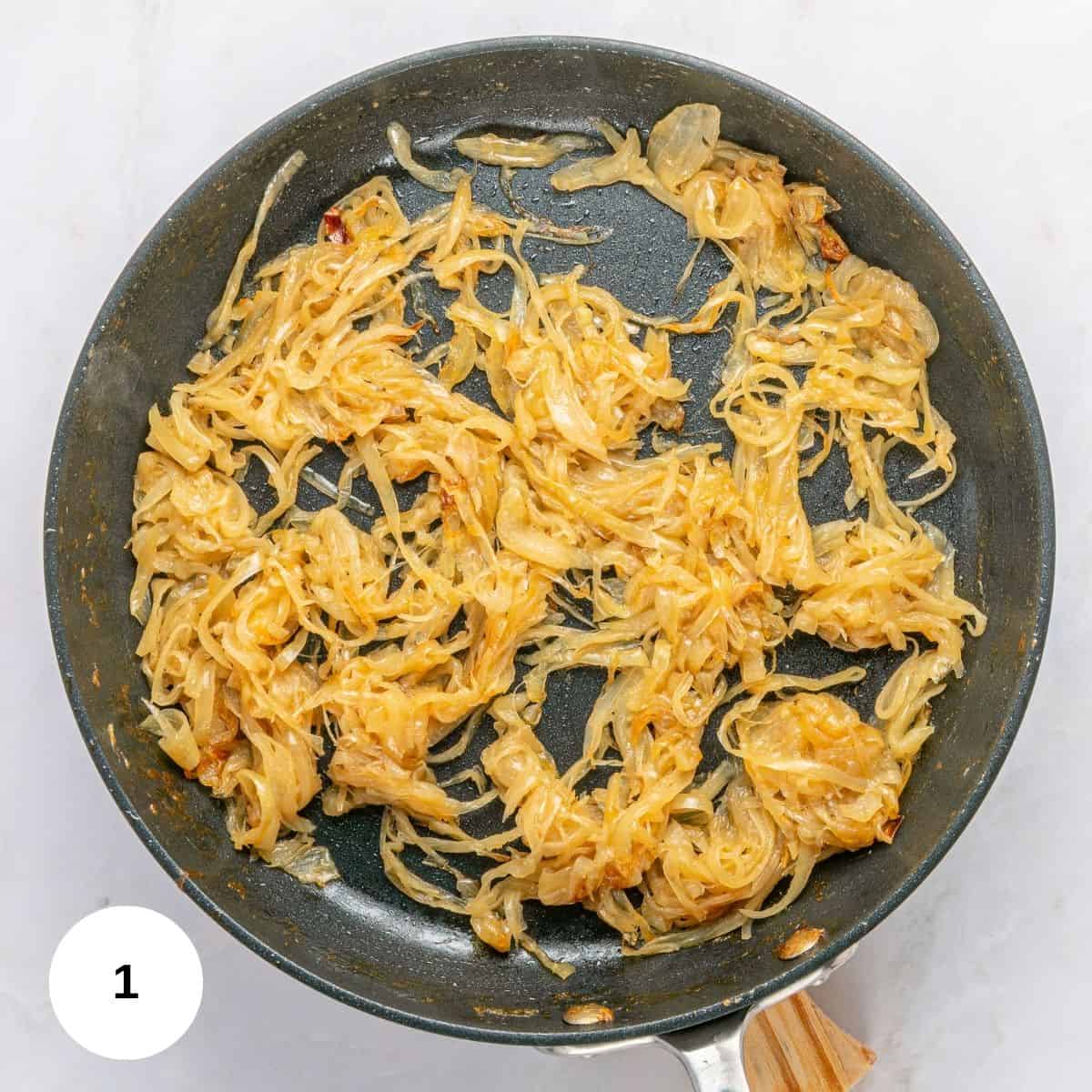 A nonstick frying pan filled with golden-brown caramelized onions, perfect for French Onion Pasta, sits on a light-colored surface. A white circle with the number 1 is in the lower left corner of the image.