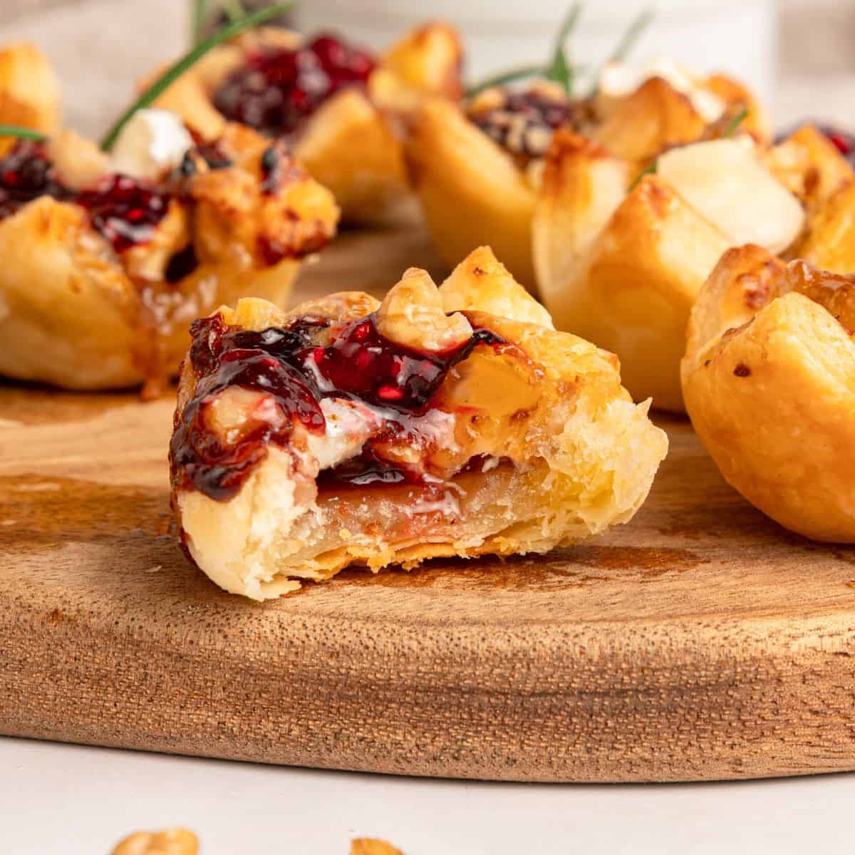 Close-up of flaky Puff Pastry Bites filled with melted brie cheese and cranberry jam, garnished with nuts on a wooden board. One cup in the foreground has a bite taken out, revealing the creamy filling inside.
