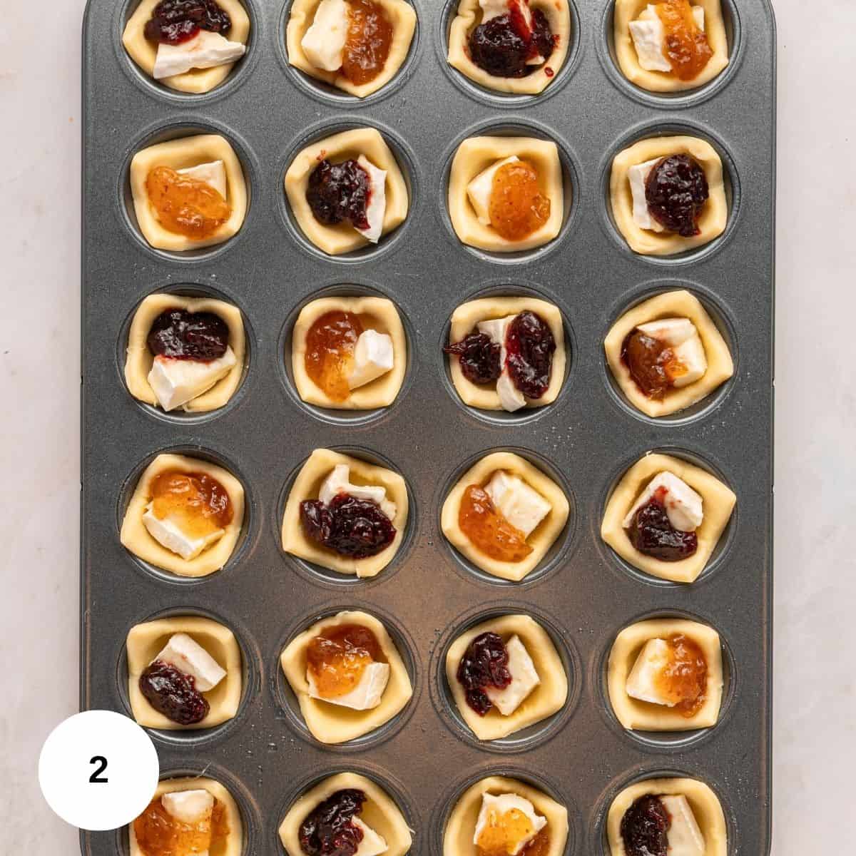 A muffin tin filled with unbaked pastry cups, each holding creamy Brie cheese and either cranberry or orange fruit jam, arranged in neat rows on a light surface.