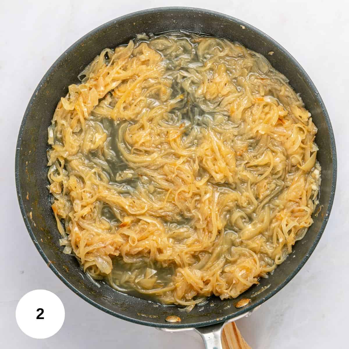 A skillet filled with caramelized onions cooking in a light brown liquid, perfect for French Onion Pasta, shown from above on a white surface. A small circle with the number 2 is in the bottom left corner.