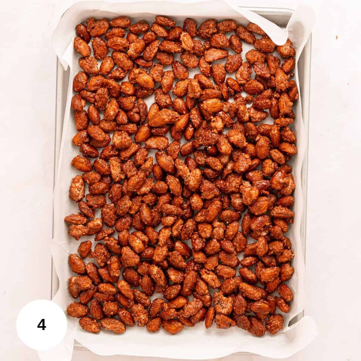 A baking sheet lined with parchment paper holds a single layer of cinnamon roasted almonds, sugar-coated and golden brown. The nuts are evenly spread out, with a white circle bearing the number 4 in the bottom left corner.