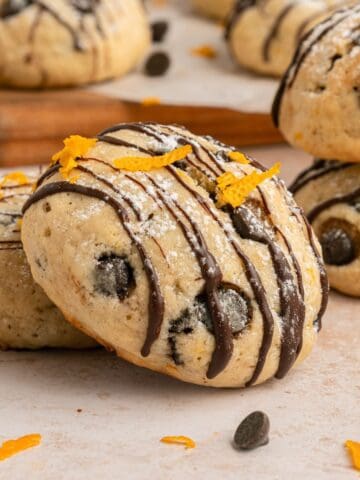 Close-up of Cannoli Cookies with chocolate chips, drizzled with chocolate and sprinkled with orange zest, with more cookies and chocolate chips in the background on a light surface.