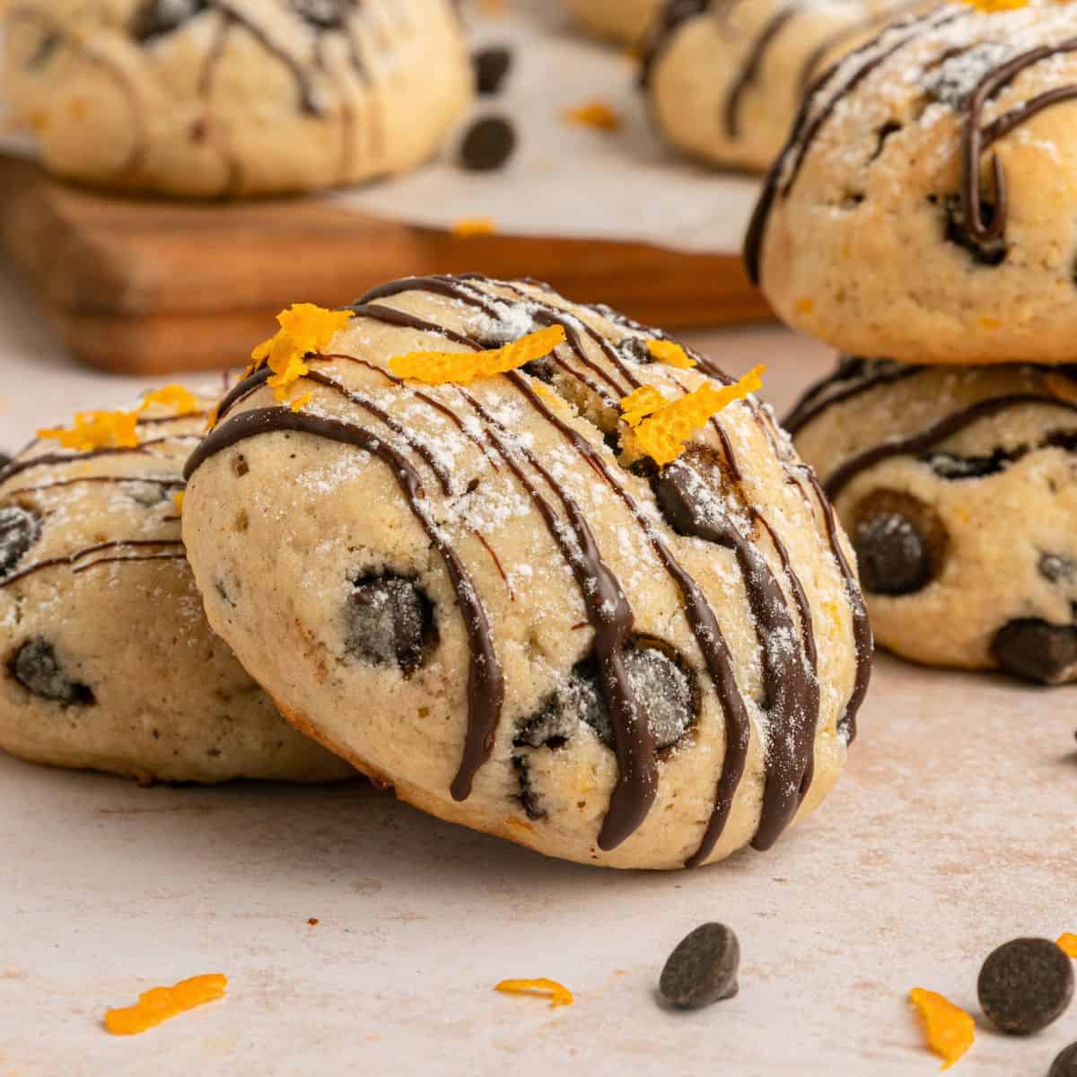 Close-up of Cannoli Cookies with chocolate chips, drizzled with chocolate and sprinkled with orange zest, with more cookies and chocolate chips in the background on a light surface.