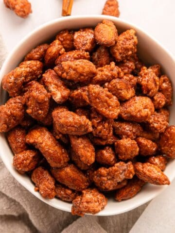 A white bowl filled with cinnamon roasted almonds, coated in a shiny caramelized sugar layer, sits on a light-colored cloth on a white surface.