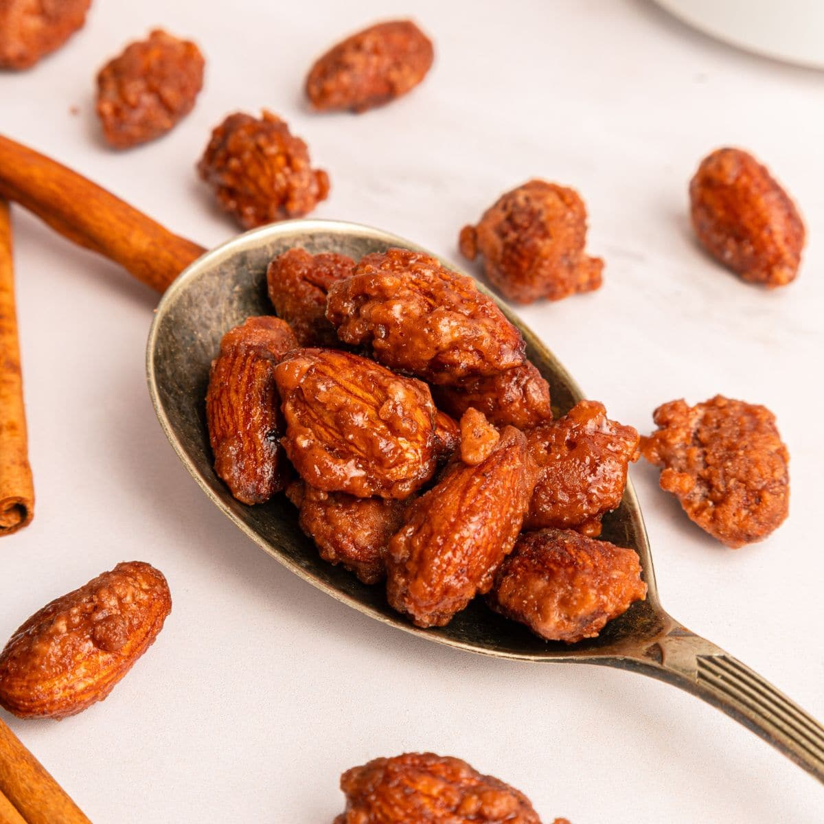 A spoon filled with sugar-coated cinnamon roasted almonds rests on a white surface, with more candied almonds and cinnamon sticks scattered around.