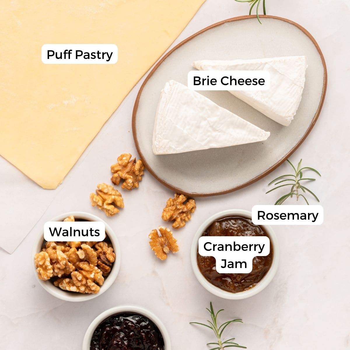 A flat lay for Cranberry Brie Puff Pastry Bites: labeled ingredients on a white surface include a sheet of puff pastry, brie wedges on a plate, bowls of walnuts and cranberry jam, and sprigs of rosemary.