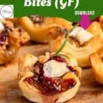Close-up of Gluten-Free Cranberry Brie Puff Pastry Bites garnished with rosemary on a wooden board. Text at the top reads Cranberry Brie Bites (GF) with a download button and a small circular logo in the corner.