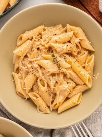 A beige bowl filled with creamy French Onion Pasta, topped with shredded Gruyère cheese, black pepper, and a sprig of thyme. A fork, a gray napkin, and fresh garlic are nearby.