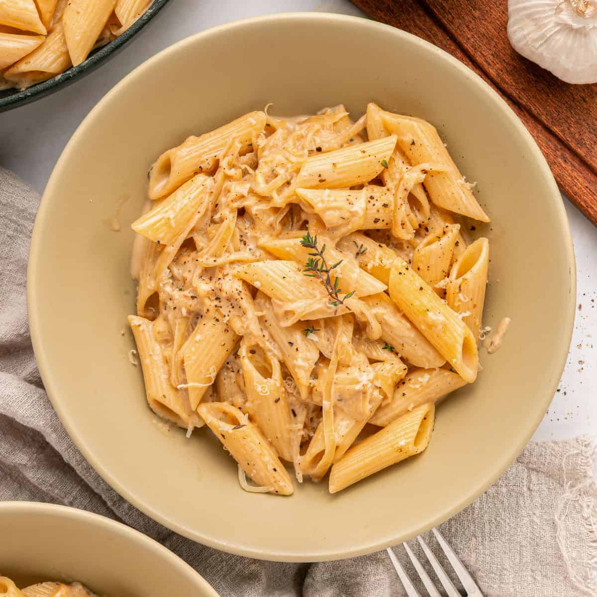 A beige bowl filled with creamy French Onion Pasta, topped with shredded Gruyère cheese, black pepper, and a sprig of thyme. A fork, a gray napkin, and fresh garlic are nearby.