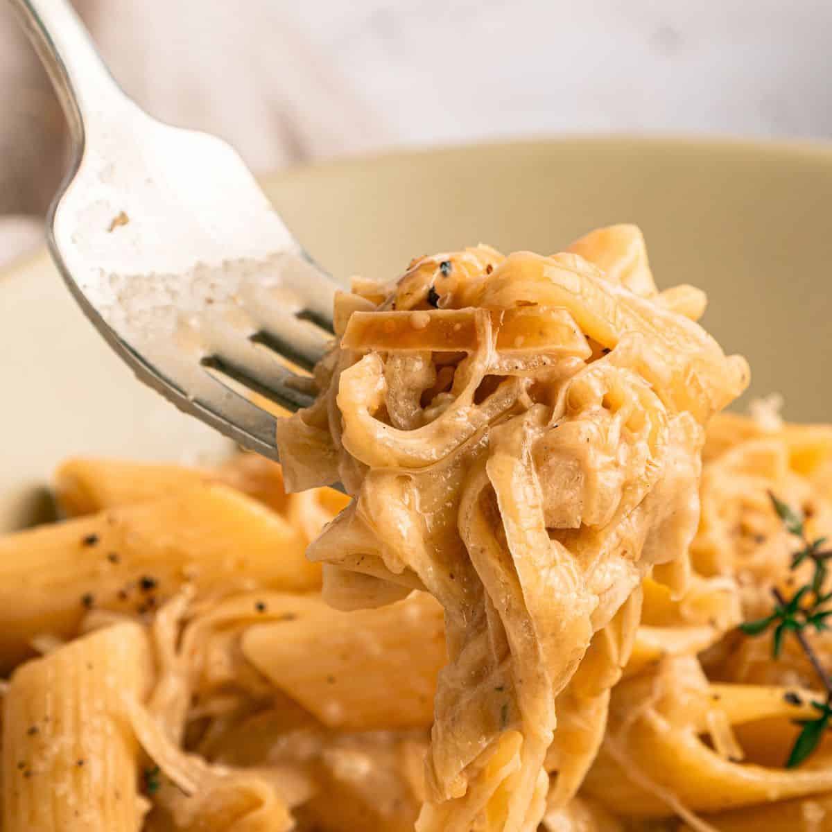 Close-up of a fork holding creamy French Onion Pasta, with penne and fettuccine coated in a rich Gruyère sauce. The dish is garnished with black pepper and herbs in a beige bowl.
