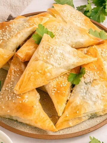 A plate of golden, flaky Greek Spanakopita Triangles topped with sesame seeds and garnished with fresh parsley. The pastries are arranged neatly on a round plate, with a small dish of dipping sauce nearby.