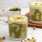 Two glasses of green pistachio latte, made using a delicious pistachio latte recipe, are topped with foam and chopped pistachios on a white surface, surrounded by scattered pistachios and a small bowl of green sauce.
