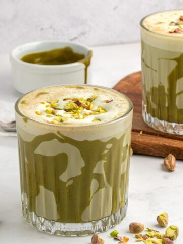 Two glasses of green pistachio latte, made using a delicious pistachio latte recipe, are topped with foam and chopped pistachios on a white surface, surrounded by scattered pistachios and a small bowl of green sauce.