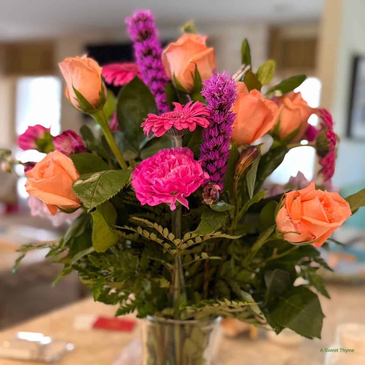 A Sunday Thymes bouquet of orange roses, bright pink flowers, and purple spikes arranged with green leaves in a glass vase sits elegantly on a tabletop indoors.