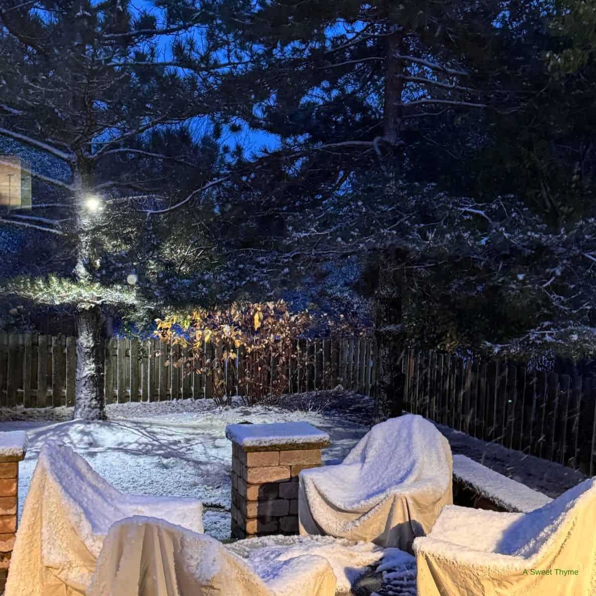A backyard patio at dusk, lightly dusted with snow. Outdoor furniture is covered and surrounded by trees and a wooden fence. Warm light, reminiscent of Sunday Thymes, illuminates part of the yard, creating a peaceful winter scene.
