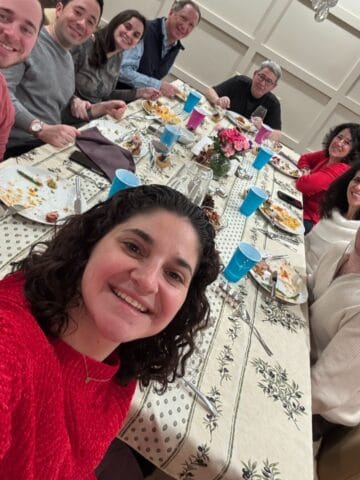 A group of nine people sit around a dining table set with plates, food, and drinks, smiling for a Sunday Thymes photo. A woman in red takes the selfie, capturing the cheerful and festive atmosphere.