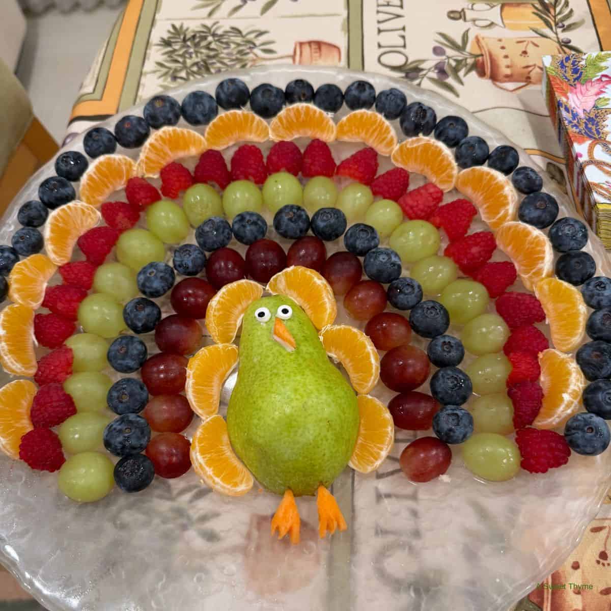 A colorful fruit arrangement shaped like a peacock, crafted by Sunday Thymes, features a pear body, orange slice feathers, and rows of blueberries, raspberries, grapes, and tangerines forming a vibrant fan behind it.