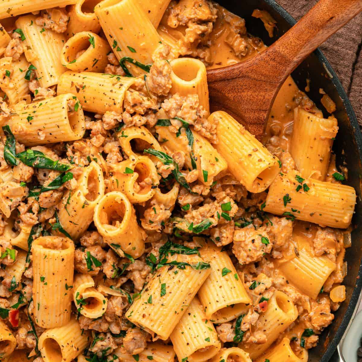 A close-up of creamy pasta with Italian sausage in a rich sauce, fresh herbs, and rigatoni stirred together in a skillet with a wooden spoon.