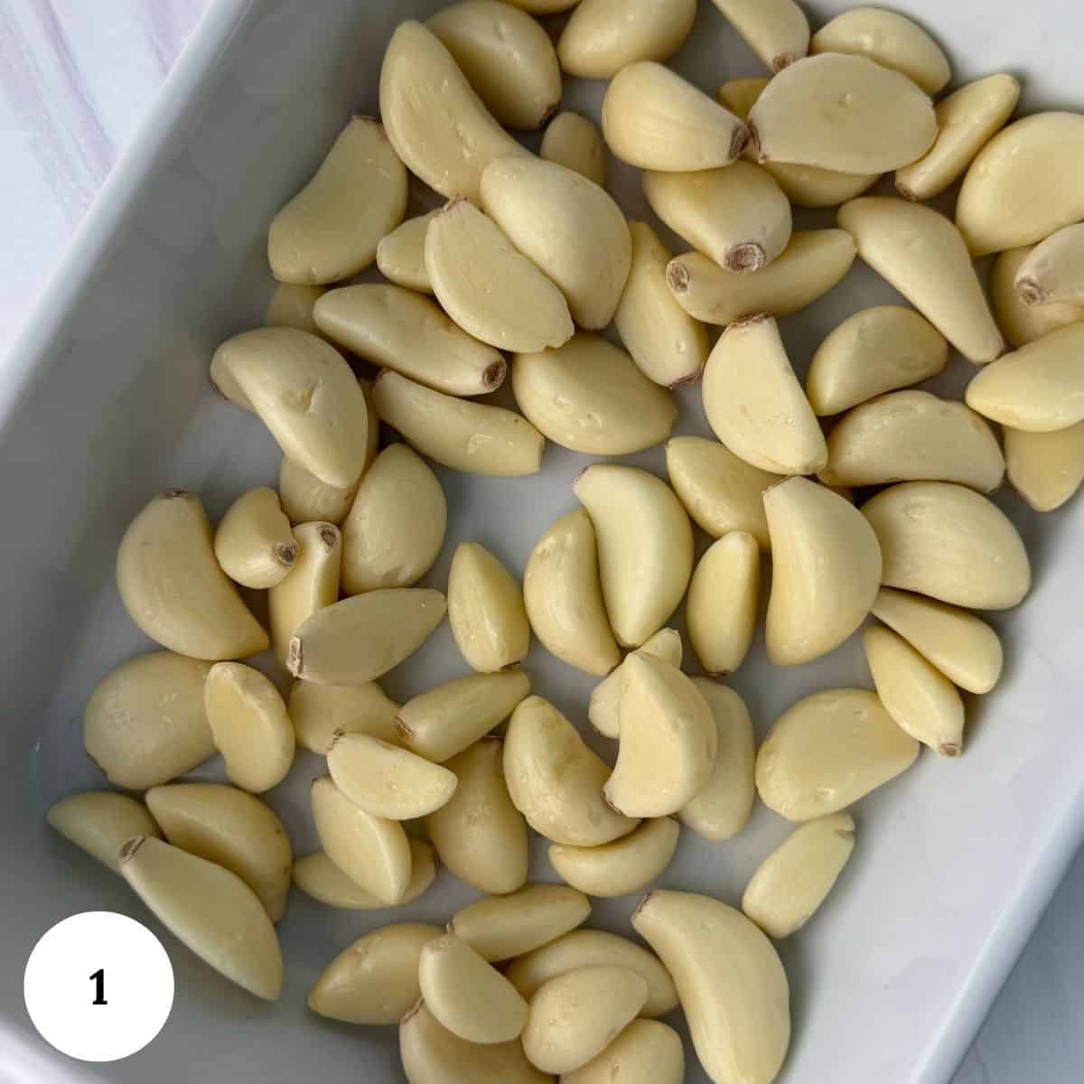 A white dish filled with peeled garlic cloves, perfect for making garlic confit, is spread out evenly. A small white circle with the number 1 is in the bottom left corner of the image.