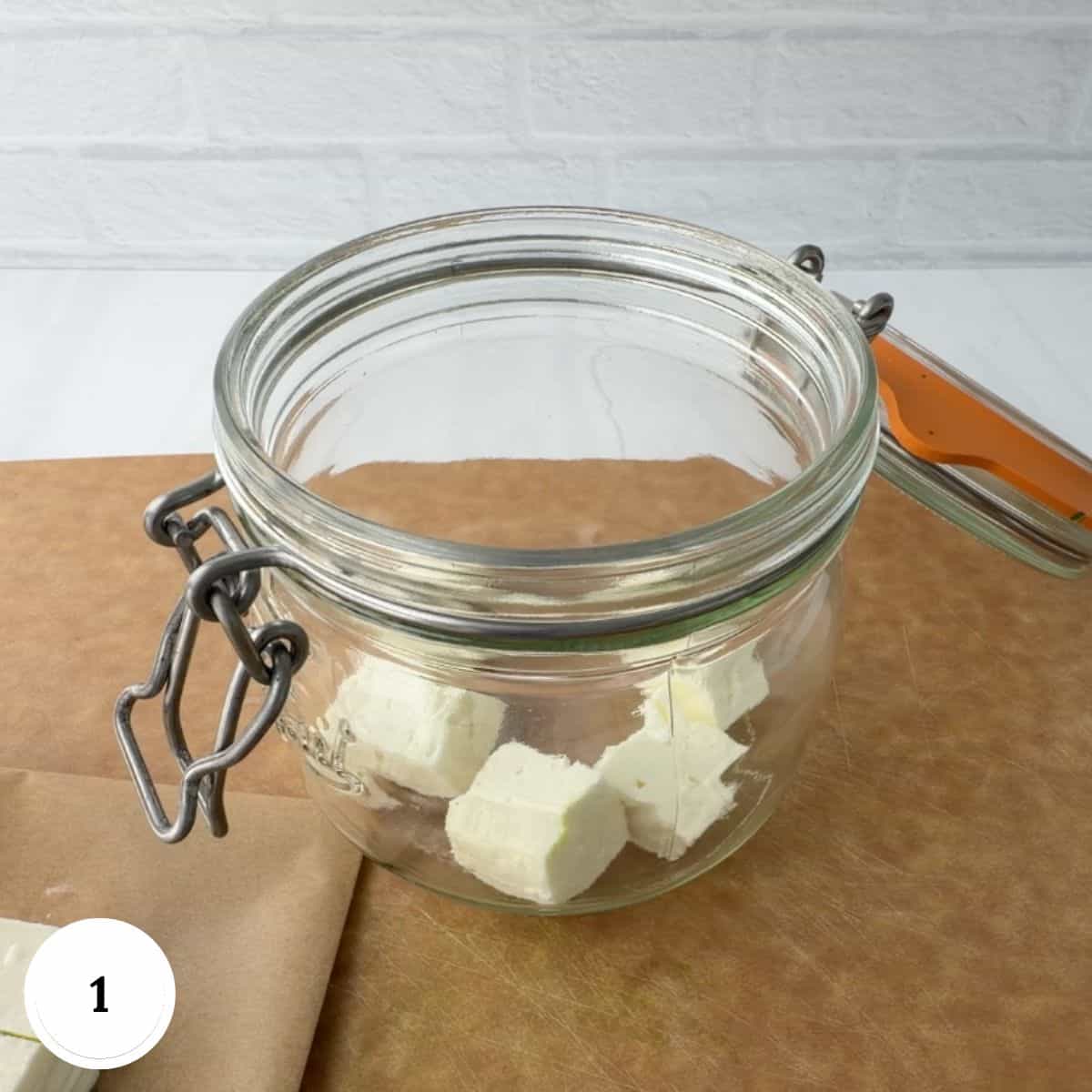 An open glass jar sits on a brown surface, partially filled with several cubes of marinated feta cheese, with more cheese cubes on a piece of parchment paper beside the jar.