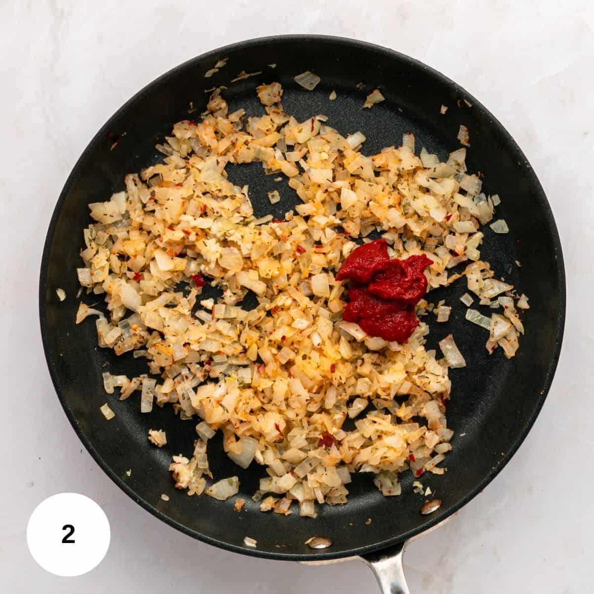 Chopped onions and seasonings are sautéing in a black skillet; a small amount of tomato paste is added to one side, forming the flavor base for Italian Sausage Pasta. The pan rests on a white surface with a white circle marked 2 in the bottom left corner.