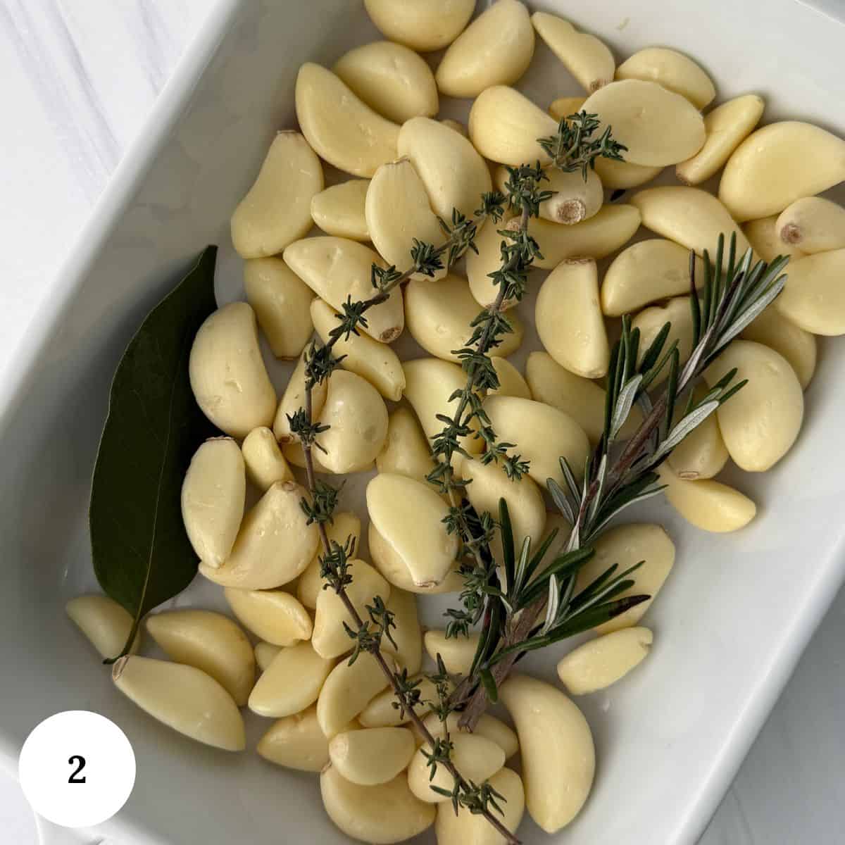Peeled garlic cloves in a white dish, prepared for garlic confit, are garnished with a bay leaf, thyme, and rosemary. A white circle with the number 2 appears in the bottom left corner.
