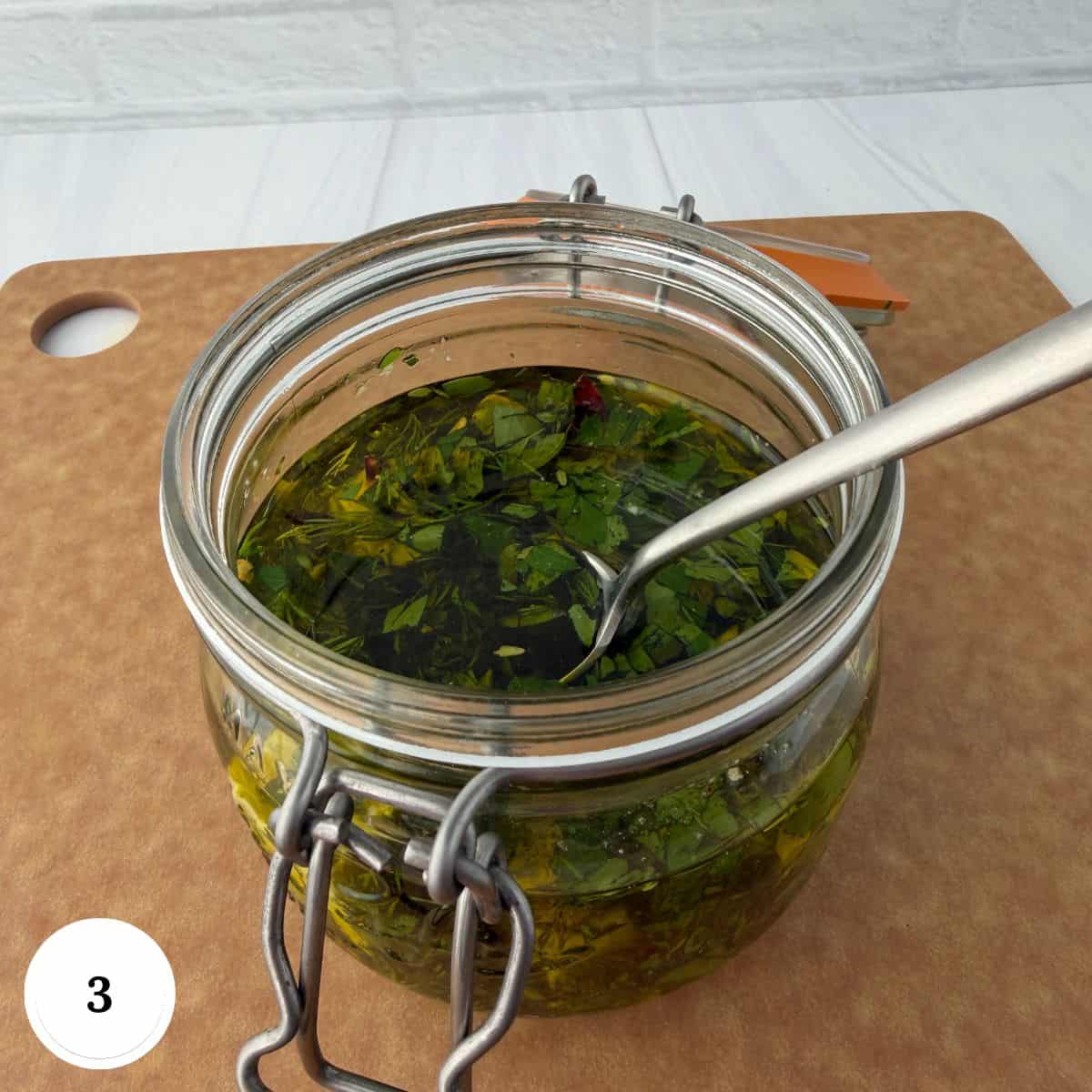 A glass jar filled with chopped herbs, oil, and marinated feta cheese sits on a brown cutting board, with a metal spoon inside. The jar has a hinged lid, against a white brick wall. A circle with the number 3 is in the corner.